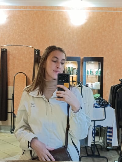 A young woman is taking a selfie in what appears to be a clothing store or boutique. She is wearing a light-colored jacket, a turtleneck sweater, and has a purse with a shoulder strap. The reflection shows clothing racks with garments, glass display cases filled with products, and orange textured walls. The location is Verkh-Sosnova, Russia.