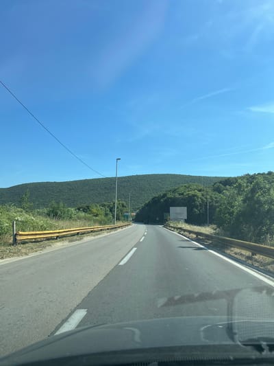 The image shows a view from the perspective of someone driving on a road. The road is paved and has white lane markings. There are yellow guardrails on either side of the road. The road curves to the right in the distance. To the left of the road, there are grassy areas and trees, with a power line stretching diagonally across the sky. To the right, there is a white billboard with the text "CAROLEA" and "12 km" and an arrow pointing to the right. Beyond the road and the billboard, there are densely vegetated, rolling hills. The sky is a clear blue with a few wispy clouds. The scene appears to be a scenic route in a rural or semi-rural area. The provided location "Bratica - Braticë, Montenegro" is consistent with a region that features such landscapes.