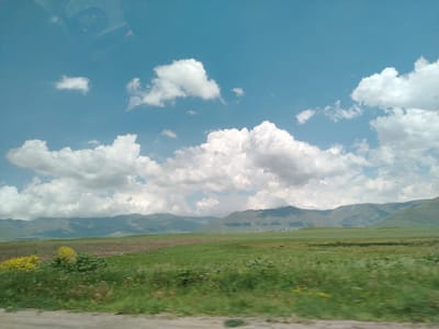 The image depicts a vast, open landscape with rolling hills in the background under a bright blue sky dotted with fluffy white clouds. The foreground features a green meadow interspersed with patches of brown soil, suggesting agricultural fields. In the mid-ground, the terrain continues with more green fields and a distant village or cluster of buildings nestled at the foot of the hills. Yellow wildflowers are visible in the left portion of the frame. The overall impression is one of rural tranquility and natural beauty. The scene is consistent with the landscape found in and around Mirak, Armenia, which is known for its mountainous terrain and agricultural areas.