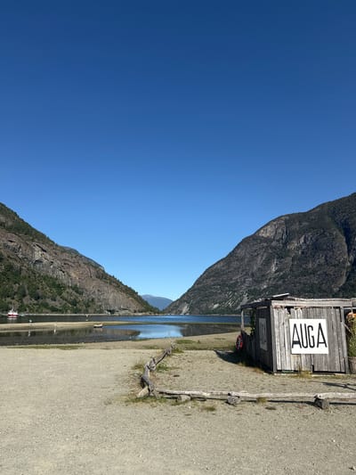 In Lærdalsøyri, Norway, a clear blue sky stretches over a tranquil body of water framed by steep, tree-covered mountains. A sandy shoreline leads to the water's edge, where a small wooden structure with "AUGA" written in large black letters on its side stands. A red life preserver hangs on the side of the structure. In the distance, a red and white boat is moored, and a few small figures are visible on the shore and in the water. The scene evokes a sense of peaceful isolation in a natural landscape.