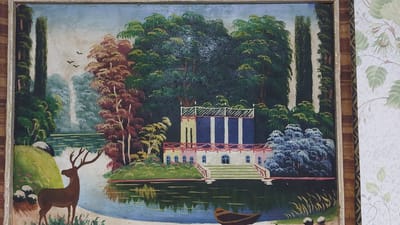 The image is a painting depicting a pastoral landscape with a building and a deer. In the foreground, a brown deer with antlers stands on a grassy bank. To the left of the deer, there are some white rounded shapes, possibly sheep or rocks, and a large grey rock. Behind the deer, a body of water reflects the surrounding scenery.
The middle ground features a building with a prominent blue and white facade, flanked by decorative red railings. The building has a series of arched windows on the lower level and appears to have a balcony or terrace on the upper level with white railings. Steps lead down from the building to the water. Several trees with red and yellow foliage are visible to the left of the building, and a dense dark green forest forms the backdrop. Tall, slender trees, possibly cypresses, frame the scene on either side. Three birds are depicted in flight in the sky above the water. A small, dark-colored boat is partially visible in the water in the lower right corner.
The painting is framed by a wooden border. The background of the photograph shows a portion of wallpaper with a floral pattern.
The image does not provide any information that would allow for the identification of a specific location in the city of Oleśnica, Poland.