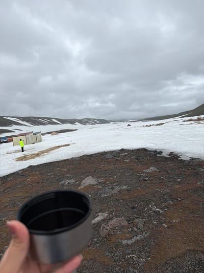 The image depicts a bleak, overcast day in a snowy, mountainous landscape, likely near Petropavlovsk-Kamchatskiy, Russia. The foreground is dominated by rocky, reddish-brown terrain with sparse vegetation and scattered white bone fragments. In the middle ground, a person wearing a bright yellow jacket stands next to a row of small, prefabricated buildings, one of which is painted with a maroon roof. Several vehicles are parked behind the buildings. A vast expanse of snow covers the undulating terrain stretching into the distance, with hills or mountains on either side. The sky is filled with heavy, gray clouds. In the immediate foreground, a hand holds a silver and black thermos lid.