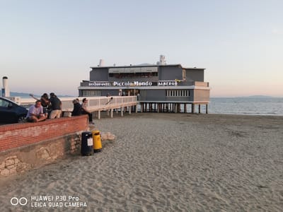 A restaurant and hotel named "Ristorante Piccolo Mondo Albergo" is situated on stilts over the water in Follonica, Italy. The building is painted gray with white accents and features prominent signage. A wooden pier connects the building to the sandy beach where two yellow and blue trash cans are visible. On the beach, four individuals are seated on a low brick wall. One person is taking a selfie with two others. In the background, the sea stretches to the horizon under a pale sky. The scene appears to be during late afternoon or early evening.