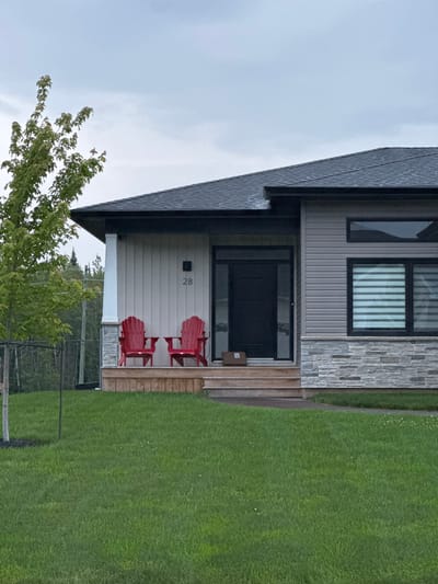 The image shows the exterior of a modern house, likely located in Dieppe, Canada, on an overcast day. In the foreground, a well-maintained green lawn stretches across the scene. A young tree with light green leaves stands to the left of the house. The house features a dark grey roof, a light grey vertical siding on the upper portion, and a stone facade on the lower portion. A dark grey front door is centrally located under a covered porch. Two bright red Adirondack chairs are placed on the wooden porch. Above the door, the house number "28" is visible. A small cardboard box rests on the bottom step of the porch. To the right of the entrance, a large window with horizontal blinds is present. The background reveals a dense treeline.