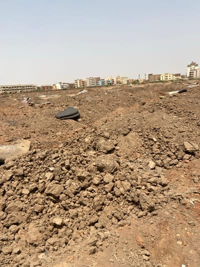 The image shows a large dirt field in Khartoum, Sudan, at the coordinates 15.528, 32.571. The field is covered in loose, brown soil and rock, with a few pieces of debris scattered throughout. There are buildings in the distance. The sky is clear and blue. There are no people visible in the image.