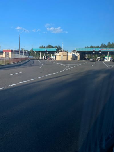 The image depicts a border crossing point, likely in Belarus, given the location context. The scene is set outdoors under a clear blue sky with scattered white clouds, suggesting it's daytime. The weather appears to be pleasant, with good visibility.
The primary focus is on a road leading towards a border checkpoint structure. The checkpoint building has a teal-colored roof and features several lanes with what appear to be booths for processing vehicles and individuals. Some vehicles, including cars and vans, are visible at the checkpoint, suggesting ongoing traffic or waiting. A few people are present, likely officials or travelers interacting at the border.
The road itself is asphalt, marked with white lines. To the left of the road, there are buildings and fencing, with trees visible in the background behind the checkpoint structure, indicating a somewhat rural or semi-urban setting. Streetlights are also present along the roadside.
Text is visible on the teal-colored structure, which reads "БЕРАСТАВІЦА" in Cyrillic script, confirming the location as Berastavitsa, a Belarusian border crossing. The overall atmosphere seems orderly and functional, characteristic of a border control area. The time of day is likely morning or afternoon due to the bright sunlight casting shadows.