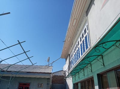 The image presents an exterior view of two residential or commercial structures in Kasan, Uzbekistan, under a clear, bright blue sky. On the right, a two-story building with a light gray-green textured facade features a long, multi-paned upper window with white frames, strongly reflecting the sky. Below this, a teal-colored, translucent corrugated plastic awning, supported by six ornate black wrought iron scrollwork brackets, extends over lower windows with brown frames. The upper facade includes a decorative light beige and white striped fascia board with elongated oval patterns, and a white satellite dish is mounted high on this wall. A red and white geometric pattern is visible on the upper right corner of the building. To the left, a lower, single-story building or annex with a corrugated metal roof is visible. A skeletal metal framework of horizontal and diagonal bars, possibly a trellis or antenna support, extends above this roof. Another white satellite dish is affixed to the roof of this lower structure. A red door and a framed object (possibly artwork or a sign) are partially visible on the lower building's wall. A section of a light brown brick wall is seen in the background between the two buildings. The scene is devoid of people, actions, or interactions.