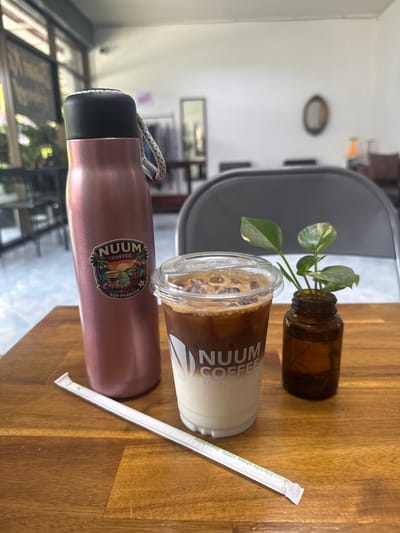 The image depicts a table setting with a bottle, an iced coffee, a straw, and a water bottle. The iced coffee is in a clear plastic cup with a lid, labeled "NUUM COFFEE KOH PHANGAN". The straw is white and labeled "Eco Friendly Straw 100% degradable". The water bottle is pink and also labeled "NUUM COFFEE KOH PHANGAN". The bottle is brown and contains a small plant. The table is made of wood. In the background, a chair and a window are visible. This scene most likely takes place at a NUUM Coffee location in Baan Kai, Thailand.