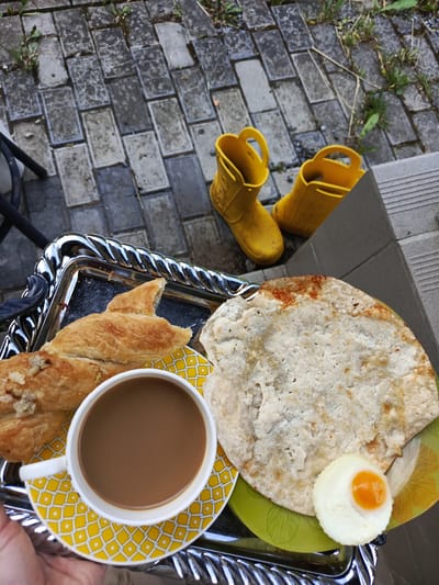 An overhead, close-up shot shows a silver tray with a breakfast of coffee, pastry, and a flatbread with a small, round, hard-boiled egg on a green plate. The tray sits on a cobblestone patio outside. In the background, a pair of bright yellow rubber boots are placed on the patio. The scene appears to be in Alexandrovskaya, Russia, suggesting a casual outdoor meal setting.
