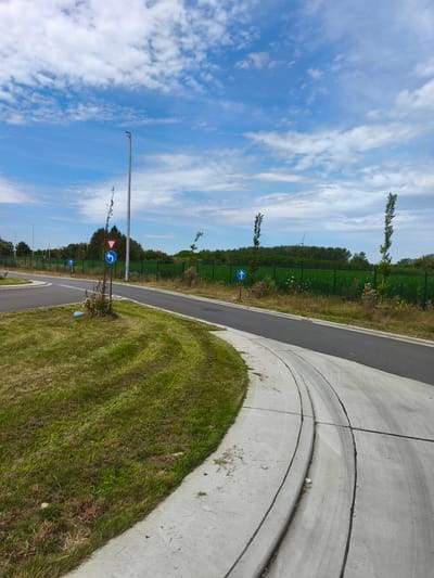 This is a photograph taken in Diepenbeek, Belgium, depicting a paved pathway with a concrete curb in the foreground, curving to the left. The pathway is bordered by a grassy area on the left, which has some dry patches. In the background, there is a dark asphalt road running parallel to the pathway. Beyond the road is a green metal fence, and behind that, a field of tall green crops. A row of trees is visible further in the distance, silhouetted against a bright blue sky with scattered white clouds. Several blue circular traffic signs with white arrows and a red triangular yield sign are posted on poles along the side of the road and pathway. A tall, modern street lamp stands on the left side of the frame, near a small, thin tree with dry branches. There are no people or animals visible in the image. The overall impression is one of a rural or suburban area with infrastructure for pedestrian or bicycle traffic alongside a road.