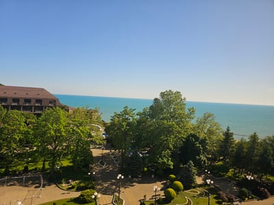 The image depicts a serene, sunlit scene overlooking a body of water, likely the Black Sea, given the location context of Lermontovo, Russia. The time of day appears to be late morning or early afternoon, with the bright, clear sky and strong sunlight casting distinct shadows. The weather is fair and warm, contributing to the tranquil atmosphere.
In the foreground, a paved walkway with a decorative pattern winds through a well-maintained park. Several lampposts with multiple globe lights are strategically placed along the path, and benches are visible, suggesting a public recreational area. Lush green grass and meticulously trimmed bushes, some shaped into rounded mounds, add to the park's aesthetic. A few trees, including large deciduous ones with full canopies and some evergreen conifers, provide shade. In the lower left of the frame, a section of temporary fencing is present, possibly indicating ongoing construction or an event setup.
Further back, a large, multi-story building with a dark, sloped roof and visible air conditioning units is situated on a slight incline. It appears to be a hotel or resort, with its architecture suggesting a tourist accommodation. Beyond the park and the building, the expansive turquoise water stretches to the horizon, meeting a clear, bright blue sky. The gentle ripple of the water indicates calm conditions.
The scene is devoid of prominent human activity, with only a few indistinct figures visible in the distance within the park. The overall impression is one of peace and natural beauty, a typical holiday resort setting bathed in pleasant weather. No visible text is present in the image.
