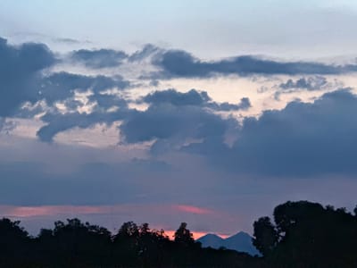 A photograph captures a twilight scene in El Bajío, Venezuela, silhouetting trees and mountains against a cloudy sky tinged with faint pink at the horizon. The sky is the most prominent feature, with layered clouds ranging from light gray to dark blue. Silhouetted in the foreground are trees and, in the distance, a pair of mountains. The overall light suggests either dusk or dawn.