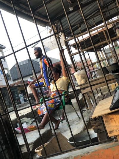 Two people sit outside, seen through metal bars. One wears a blue and white striped shirt, the other a yellow shirt and colorful skirt. They're on a concrete surface with a green chair nearby. Background features buildings, objects covered with white cloths, and overhead metal structures. The scene is viewed at an angle through the bars.