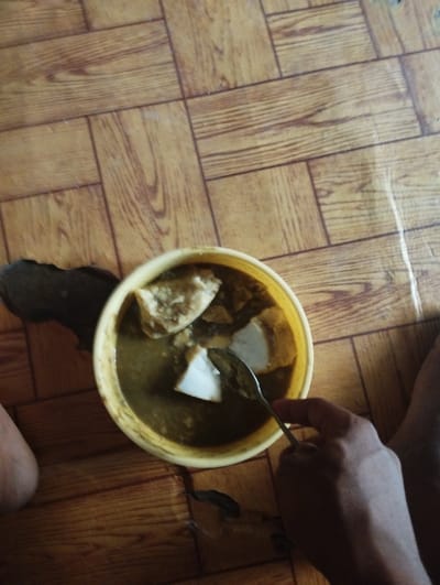 A person is sitting on the floor, using a metal spoon to eat a bowl of dark, viscous stew with white chunks of food.  The bowl is yellow and has a brown rim. The person's right hand is visible, holding the spoon, and their left foot is partially visible on the left side of the image. The floor is made of light brown square tiles with darker brown lines separating them.  The scene is likely in a home in Bajoga, Nigeria, based on the given coordinates.