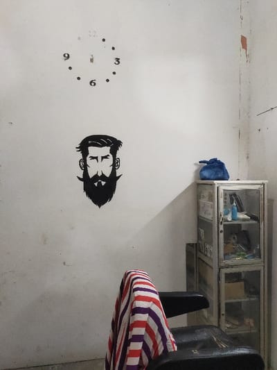 The image displays the interior of a room, likely a barbershop, located in the city of Tanjong, Indonesia. The walls are an off-white color, exhibiting signs of wear, dirt, and some peeling paint, particularly along the right corner. Mounted on the upper left wall is an incomplete clock face with black numerals '9', '6', and '3', along with dots marking other hours; the clock mechanism is visible but no hands are present. Below the clock, a black, stylized decal of a bearded male head with a mustache is affixed to the wall. To the right, a two-tiered, metallic-framed display cabinet with glass doors stands against the wall. The upper compartment contains various items, including a blue spray bottle and other containers. The lower compartment holds less distinct objects. A crumpled blue drawstring bag rests on top of this cabinet. In the foreground, a black chair, possibly a barber chair, is partially visible with its back facing the viewer, draped with a vertically striped cloth in red, white, and purple/blue. A sharp shadow is cast diagonally across the right wall and a section of the main wall. No individuals are present in the scene.