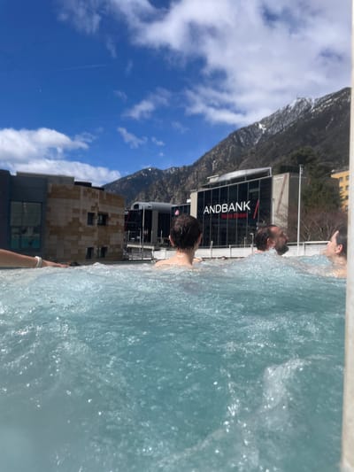 The image depicts a daytime scene from the city of Les Escaldes, Andorra. Three individuals are partially submerged in what appears to be a pool with light blue water. The individuals' faces and heads are visible above the water's surface. Behind the pool, the Andbank building is identifiable by the large lettering on its exterior. A mountain rises in the background, partially obscured by the building. The sky is blue with scattered white clouds.