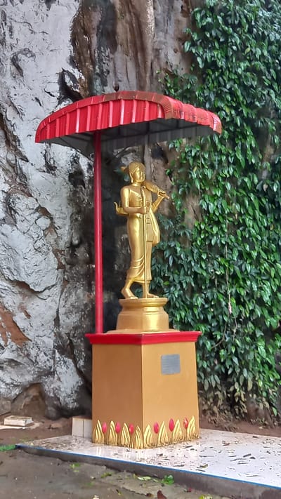 A golden statue of a robed figure carrying a staff stands atop a decorated base beneath a red umbrella-like structure, set against a background of rock and lush green vegetation. The statue and its stand are brightly lit, while the surrounding environment is partially shaded. The location is near a road and within the city of Ban Pak Ra, Thailand.