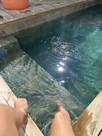 A sunlit swimming pool is pictured from an overhead perspective. The pool has a stepped entrance. The water reflects sunlight, creating a sparkling effect. Two human feet are dipped into the pool, near the edge of a wooden deck. An orange cushion is visible to the left of the pool. The location is La Couarde-sur-Mer, France.