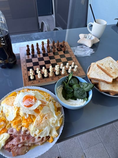 The image depicts a breakfast spread laid out on a gray, reflective table, likely outdoors given the lighting and surrounding environment which suggests a terrace or patio. On the table, a chessboard with pieces set up for a game is positioned centrally. To the left of the chessboard is a large black bottle of soda. To the right of the chessboard, there is a white ceramic mug with a black straw, next to a small, star-shaped decorative dish. Further to the right, a bowl containing toasted bread slices sits alongside a blue bowl filled with ricotta cheese and fresh spinach leaves. In the foreground, a white plate holds a breakfast of fried eggs, ham, and possibly other savory items. The overall scene suggests a casual breakfast, possibly before or during a game of chess. The location is identified as Nea Potidea, Greece. There are no people visible in the image.