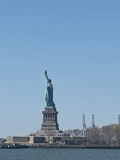 The image shows the Statue of Liberty viewed from the water, likely from a boat. The statue is green and its base is gray. Behind the statue, on the right, are trees, a building, and industrial cranes. The sky is clear and blue. The water is dark gray.