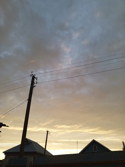 The image depicts a cloudy sky with hues of grey and yellow, suggesting either dusk or dawn. In the foreground, there is a dark fence and the rooflines of two houses. One house has a dark, pitched roof and a visible window in its gable end, painted blue. The other house, partially obscured, also has a dark pitched roof. A wooden utility pole stands prominently in the mid-ground, leaning slightly to the right, with electrical wires stretching horizontally across the frame. Another smaller utility pole is visible to the right of the first house. The overall scene is located in the city of Buraly, Russia.