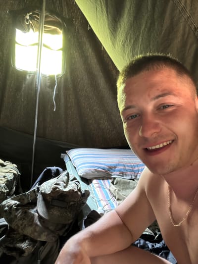 The image presents a close-up, rotated portrait of a young man inside a tent or temporary shelter. The man, who appears to be in his late teens or early twenties, has very short, light-colored hair and a fair complexion. He is smiling widely, showing his teeth, and looks directly towards the camera, conveying a friendly and content expression. He is bare-chested and wears a thin gold chain around his neck.

The interior of the shelter is made of dark green or olive drab fabric. The scene is illuminated by a bright, rectangular artificial light fixture, possibly an LED panel, hanging from a metal pole, indicating that the photograph was taken in a dark environment, likely at night.

To the right of the man, a pillow with blue, white, and red stripes is visible. Below the pillow, a pile of gear is present, including a camouflage-patterned hat or piece of clothing, suggesting military or outdoor adventure equipment. Another bag or backpack is also part of this gear. The setting, combined with the visible gear, suggests a military, camping, or field environment, consistent with the provided location context of Ostar Kamak, Bulgaria. There is no visible text in the image.