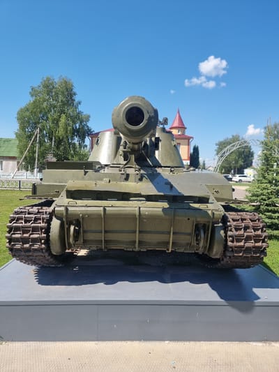 The image shows a military tank, likely a monument, displayed outdoors on a sunny day with a clear blue sky and scattered clouds. The tank is olive green and is positioned on a grey, rectangular platform. The front of the tank, including its prominent gun barrel, is the main focus. The tank's tracks are visible on both sides. In the background, there are trees, a building with a red-tiled roof and a tower, a white fence, and what appears to be a decorative archway structure. The overall setting suggests a public space, possibly a park or memorial in the city of Атяшево, Russia, dedicated to military history. There are no people visible in the image.