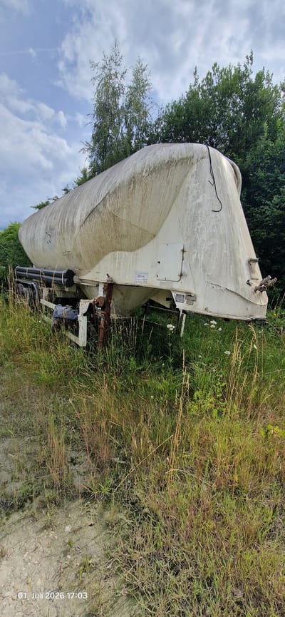 A large, white, cylindrical tank, possibly a cement mixer or silo, is the primary object in this image, lying on its side in a grassy, overgrown area. The tank appears weathered and dirty, with streaks and stains on its surface. It is situated outdoors, with a backdrop of lush green trees and a cloudy blue sky.
The tank is resting on what appears to be a rusted metal frame or chassis, suggesting it might be a trailer or part of a larger vehicle. Some black pipes or hoses are visible attached to the underside of the tank. The surrounding area is filled with tall, dry grass and some smaller white flowers.
The image is oriented sideways, with the sky and trees appearing on the left and the ground on the right. There is a timestamp in the lower right corner which reads "01. Juli 2026 17:03". The lighting suggests it is late afternoon, and the weather appears to be partly cloudy.
The scene evokes a sense of abandonment or disuse, with the vehicle lying off the road in a natural, untamed environment. There are no people visible in the image. A label is attached to the side of the tank with some German text, but it is too small to be legible. The overall impression is one of a forgotten piece of industrial equipment slowly being reclaimed by nature.