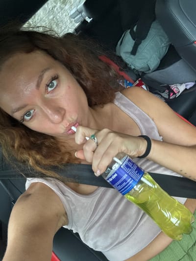A woman is in a car, wearing a light pink top and a seatbelt. She is holding a plastic bottle filled with yellow liquid and appears to be using a white straw. The car's interior is visible, including a backpack and some clothing items. The location is stated to be Quản Bạ Commune, Vietnam.