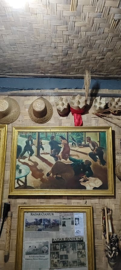 The image displays an interior scene in Cikanyere, Indonesia, characterized by traditional Indonesian architecture and artifacts. The ceiling and walls are constructed from light-colored woven bamboo panels.
Affixed to the upper wall, positioned above a horizontal bamboo beam, are five traditional straw hats. Two broad-brimmed hats are on the left, while three smaller, shaped hats are on the right, one of which is adorned with a red fabric piece. A small bundle of dried plant stalks is tucked into the wall structure near the center-right.
Centered on the wall is a large, rectangular painting enclosed in a gold-colored frame. The painting depicts a dynamic outdoor or semi-outdoor scene with approximately eight figures engaged in various actions. Figures are dressed in traditional clothing. Notable actions include a man on the left in a fighting stance, potentially wielding a stick; another man pulling a woman in a red patterned dress; and at least one figure lying on the ground in the foreground. An overturned small table with a teacup and saucer is visible on the left side of the painting. The background features trees and architectural elements, suggesting a courtyard or open-air structure. Strong shadows are cast on the ground within the painting.
Below this painting is a second, smaller gold-framed display containing multiple newspaper clippings. Visible headlines include "Radar Cianjur" and "Radar Bandung." Articles discuss subjects such as "Gunung Padang Keperintisan Zaman Old" and "Kebakaran Hutan Gunung Beser Disengaja?" (Forest Fire at Mount Beser Intentional?). Photographs within the clippings show groups of people and individual portraits.
To the left of the newspaper display, a long, dark object resembling a traditional wooden torch or staff hangs vertically. To the far right, a decorative item made of bundled natural materials is partially visible. An overhead light source illuminates the scene.