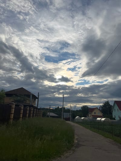 This is a vertical, medium-shot photograph taken in the city of Kostyunino-2, Russia, capturing a rural landscape under a dramatic, cloudy sky. The foreground is dominated by a dirt path winding to the right, bordered by tall, overgrown grass on the left. To the left of the path, a substantial brick and wooden house with a dark-pitched roof is visible behind a decorative brick and metal fence. Further down the path, a line of greenhouses can be seen, with several residential houses of various colors and architectural styles interspersed among trees in the background. A power line stretches diagonally across the upper portion of the frame. The sky is filled with heavy, dark grey clouds, with patches of brighter blue sky and sunlight breaking through, creating a somewhat diffused and atmospheric lighting condition. The overall scene depicts a quiet, residential area on what appears to be an overcast day.