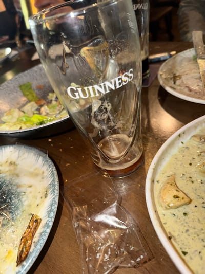 The image shows a close-up view of a table in what appears to be a restaurant setting, perhaps in Kafr Yasif, Israel. Several partially eaten plates of food are visible, including one with a creamy sauce and another with salad. An empty Guinness glass lies on its side, partially obscuring a plate of food. A transparent plastic bag is also on the table. The table surface is a medium-toned brown color. The lighting seems to be dim. No people are directly visible in the image.
