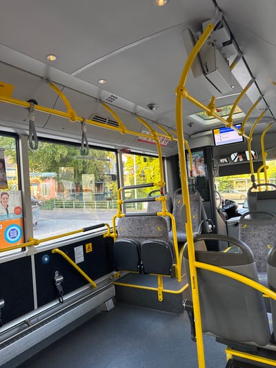 The image captures the clean and modern interior of a public bus, likely in Fehraltorf, Switzerland, during a sunny daytime. The view is from the rear of the vehicle, looking forward towards the driver's area and the front exit.
**Subjects and Objects:** The bus interior features bright yellow grab bars and poles for passengers, crisscrossing the ceiling and lining the aisle. Grey and black seating is arranged along both sides, with some seats appearing to be fixed and others potentially foldable, one bearing the text "Scouterama." The floor is dark, with patterned sections. Large windows offer views of green trees and distant buildings, indicating an outdoor, possibly suburban, environment. The ceiling has several recessed lights and ventilation grilles. Towards the front, a digital display screen is visible, along with smaller screens or mirrors and a partial view of the driver's seat. A dedicated space, likely for wheelchairs or prams, is equipped with a safety belt, a blue button, and a yellow support bar, accompanied by a wheelchair symbol. An advertisement on one window shows a person's image, a QR code, and text.
**Activity or Event:** The bus appears to be empty of passengers, suggesting it might be at a stop, between routes, or awaiting service. No specific activity by individuals is depicted.
**Notable Details:** The bright sunlight filtering through the windows indicates it is daytime with clear weather. The modern design and clean state of the interior suggest a well-maintained public transport vehicle. The presence of German text ("Willkommen" on the display and "von Zürich" on an advertisement) aligns with the provided location context of Fehraltorf, Switzerland.
**Visible Text:** Key visible text includes "827 Willkommen/W" on the overhead digital display, "Scouterama" on a seat, and various fragments from an advertisement such as "CLINIUS," "50 Min von Zürich," "re erste suchung," and "net."