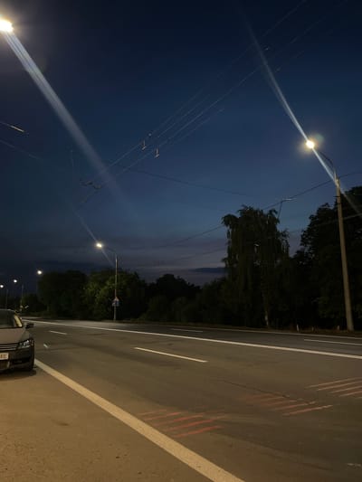 A twilight street scene in Strumivka, Ukraine features a multi-lane road with streetlights and overhead wires. A dark-colored car is parked on the left shoulder of the road. Trees line the side of the road and a blue and white road sign is visible. The sky is a deep blue with faint clouds. The road surface shows white lane markings and some faded red markings.