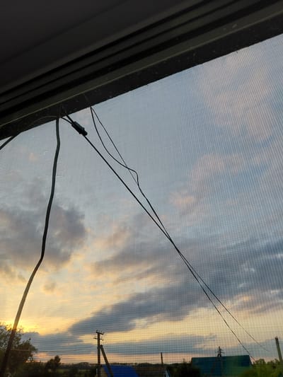 The image depicts a view through a window screen of a sunset over a rural landscape. The sky is filled with dramatic clouds illuminated by the setting sun, casting warm orange and yellow hues across the scene. In the foreground, a black cable runs diagonally across the frame, attached to a small black connector. A darker, textured frame borders the left side of the image, suggesting the window or opening through which the photograph was taken.

In the background, a telephone pole with wires is visible, along with some distant trees and structures, including a portion of a blue building. The overall impression is one of tranquility and the natural beauty of a sunset in a rural setting, possibly in Старотавларово, Russia. The time of day is clearly sunset, characterized by the low angle of the sun and the colors in the sky. The weather appears to be clear with scattered clouds. There is no visible text in the image.