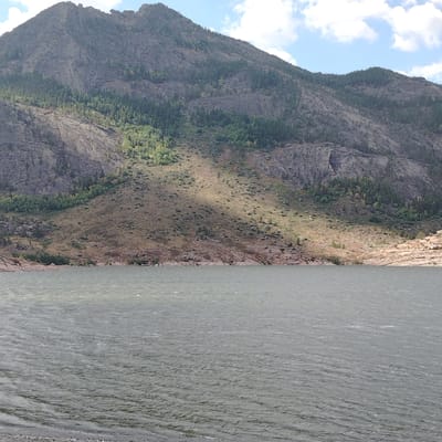 The image depicts a body of water in the foreground, with a large, rocky mountain rising behind it. The mountain is covered in sparse vegetation, with patches of trees and dry grass. The sky above is partly cloudy. The water is choppy, suggesting a windy day. The shoreline consists of rocks and pebbles. The scene appears to be a natural landscape. The location is in the vicinity of Toraygyrov, Kazakhstan, a region known for its mountainous terrain and lakes. There are no people or animals visible in the image.
