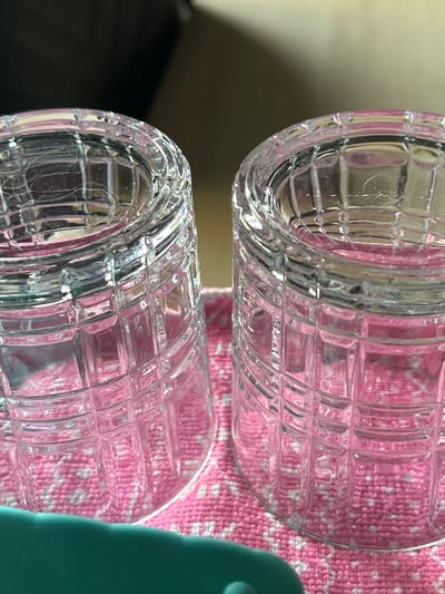 Two clear glass tumblers with a patterned texture are shown, one above the other, against a pink and white patterned fabric. The tumblers are empty and appear to be made of cut glass, with a grid-like pattern of indentations. The fabric is textured and has a light pink background with a white woven pattern. A teal object is partially visible on the right side of the image, obscuring some of the fabric and the lower tumbler. The lighting is bright, suggesting daytime, but there are no specific external cues to indicate weather or precise location within Siedlce, Poland. The focus is on the texture and transparency of the glass tumblers and the contrast with the fabric. There is no visible text in the image.