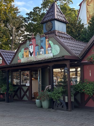 The image depicts the exterior of "Город Мастеров" (City of Masters), a craft and souvenir shop located in Yantarny, Russia. The single-story building features a brown-tiled roof with a prominent light blue clock tower topped by a darker brown pyramidal roof, displaying a circular clock face with Roman numerals. The facade beneath the clock tower is adorned with a colorful mural depicting a stylized street of various European-style houses and a black rooster against a light green background. The Russian text "Город Мастеров" is positioned below this mural.

The ground floor consists of multiple display windows showcasing merchandise, with one window clearly marked "СЕРЕБРО" (Silver) and featuring a "no smoking" symbol. An open doorway is visible, providing access to the interior. The building's base incorporates dark brown wooden cross-beams over a green paneling. The area directly in front of the building is paved with grey interlocking bricks, and several potted plants, some containing red and pink flowers, are arranged near the entrance and windows. A decorative wooden cart wheel is positioned near one of the flower boxes. Green foliage and mature trees are visible in the background under a clear sky. A portion of an adjacent building with a reddish wall, white gable, and a painted scroll design is visible on the far right. No individuals are present in the scene.