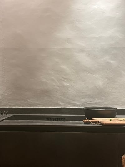 The image shows a countertop in a kitchen in Celerina/Schlarigna, Switzerland (46.514, 9.859). The countertop is made of dark grey stone. There is a black bowl with water and a wooden cutting board with green vegetables on it. There is a black metal rack on the left side of the counter and a white wall behind the counter. The countertop is illuminated with artificial light. The environment is a kitchen with a modern design. There are no people in the image.  The objects in the image are a countertop, a bowl, a cutting board, and a rack. The actions in the image are preparing food. The interactions in the image are none. The environment is a kitchen.