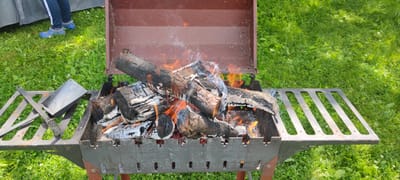 In the city of Пацейкаўскі сельскі Савет, Belarus, an outdoor scene unfolds with a portable metal grill as the central subject. The grill, a dark gray color with a reddish-brown lid, is positioned on a vibrant green grassy lawn. Inside the grill, a fire burns intensely, with logs and embers glowing orange and red, producing wisps of white smoke that curl upwards.
To the left of the grill, a person's legs are visible, clad in dark blue pants with white stripes and bright blue protective footwear. They are standing on the grass, suggesting a casual outdoor activity like a picnic or barbecue.
On the left side of the grill, a metal tool, possibly a shovel or poker, rests on a grated surface. This surface appears to be a shelf or extension of the grill. The grass surrounding the grill is short and well-maintained, with some patches of clover. The lighting suggests it is daytime, with dappled sunlight filtering through unseen trees, casting soft shadows on the grass. The overall impression is one of a preparation for or ongoing outdoor cooking event in a park or backyard setting.