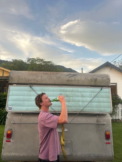 The image captures a male individual, seen in profile, actively drinking from a green glass bottle. He stands outdoors in what appears to be a residential area of Thirroul, Australia, positioned in front of a silver-grey vintage-style pop-top caravan. The man has light brown hair pulled back into a small bun and is wearing a plain mauve t-shirt. Liquid is visibly splashing from the bottle as he drinks.

The caravan features a ribbed lower section and an extended pop-top section, revealing a weathered, light blue ribbed panel. A circular white sticker with blue text reading "SAVE OUR OCEAN / SURFRIDER" is affixed to the lower left of the light blue panel. The rear of the caravan shows a red and amber taillight on the left, a reflective strip, and a horizontal metal bar. A yellow tie-down strap hangs vertically near the right side of the caravan's rear. Diagonal wires or straps secure the extended pop-top section.

In the background, to the left, a portion of a building with a yellow exterior is visible. To the right, a white house with a dark gable roof, a visible chimney stack, and a white picket fence is present. Beyond the houses, tree-covered mountains or hills rise into the distance. The sky above is partially cloudy, displaying soft light and a mix of light blue and white cloud formations. The ground in the foreground appears to be grass.