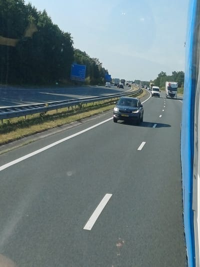 The image depicts a highway scene in Nistelrode, Netherlands. On the right side of the frame, the blue and white side of a vehicle, possibly a bus or truck, is visible. The highway itself is paved with asphalt and features white lane markings. Several vehicles are present on the highway, including a dark-colored Skoda automobile in the foreground, a white van, and several trucks further in the distance. The highway is bordered by a metal guardrail on the left, with grassy verges and trees beyond. A blue road sign is visible on the left side of the road, partially obscured by trees. The sky is clear and blue, suggesting fair weather. The overall scene is that of a typical highway environment with traffic flow.