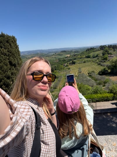 The image shows two young women in San Gimignano, Italy, overlooking a green valley under a clear blue sky. The foreground woman, who is taking the selfie, is wearing a brown and white checkered shirt, a black shoulder strap, and black sunglasses. The second woman, standing behind her, is wearing a pink baseball cap and a light green jacket, and she's holding a phone to take a picture of the valley below. The location seems to be an elevated point within or near San Gimignano, providing a wide view of the Tuscan landscape.