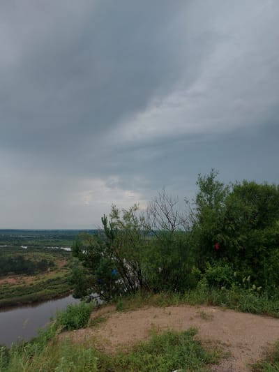 The image shows a cloudy sky over a landscape dominated by a river, lush greenery, and trees. In the foreground, a dirt path is visible on a grassy slope. The river meanders through the middle ground, bordered by dense vegetation and some open fields. The background reveals a distant horizon with more greenery and what appears to be a faint haze. A few colorful ribbons are tied to branches of the trees, one red and two blue. The scene is likely a natural area within or near the city of Parunovka, Russia, given the context provided.