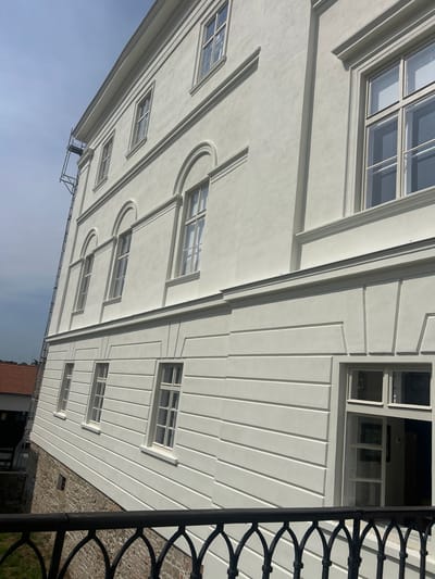 The image shows the exterior of a white building in Rosice, Czechia, with a clear sky overhead. The building appears to be a historical structure, possibly a castle or a large manor house, with multiple stories and numerous windows. The facade is decorated with architectural elements such as cornices, molding, and recessed panels. Some windows are tall and rectangular, while others are arched. Scaffolding is visible on the left side of the building, suggesting ongoing renovation or construction work. In the foreground, a black wrought-iron railing with an ornate design is partially visible. The lower part of the building is constructed with a stone foundation. To the left, a neighboring building with a red-tiled roof is partially visible. The overall scene depicts a well-maintained historical building in an outdoor setting.