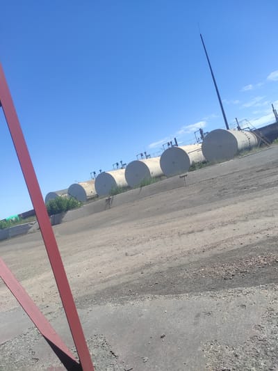 The image shows an industrial area under a clear blue sky with scattered white clouds. In the foreground, a portion of a red metal structure is visible, casting a shadow on a gravelly ground. Further back, a line of large, cylindrical storage tanks, likely for fuel or other industrial materials, are arrayed horizontally. These tanks are supported by a concrete structure and are partially overgrown with green vegetation. A tall, slender dark pole, possibly a lightning rod or communication antenna, extends from behind the tanks towards the sky. The overall scene suggests a remote industrial site within or near the city of Kyzyl, Kazakhstan, characterized by open space, industrial infrastructure, and a bright, sunny day. There are no people or animals visible in the image.
