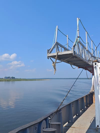 An outdoor, low-angle, medium shot captures a section of a tugboat's deck and a gangway against a bright blue sky with scattered clouds. The tugboat's dark gray hull is visible in the foreground, with a lighter gray deck stretching back. Several black bollards are present on the deck. A metal gangway, supported by beams, extends outward from the tugboat over the calm, blue water. The gangway has metal railings and is secured with ropes, one of which hangs loosely. In the distance, a faint shoreline with green vegetation and a few structures is visible, suggesting a settlement. The scene is set on the river in Pogodaevo, Russia, under a clear, sunny sky.
