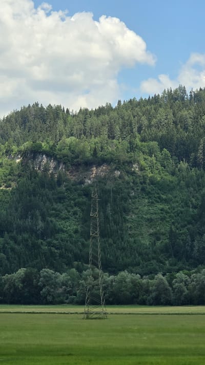 This image depicts a scenic landscape with a dense forest covering a rocky mountainside on the left. The forest is lush and green, indicating a temperate climate. In the foreground on the right, a vibrant green grassy field stretches out. A single, tall electricity pylon, constructed from metal lattice, stands prominently in the middle ground, extending from the edge of the forest into the field.
The sky above is a clear blue, dotted with fluffy white clouds, suggesting a bright and sunny day. The time of day appears to be daytime, likely late morning or afternoon, given the quality of the light. There are no visible people or animals, and no specific activity or event is taking place; the scene is a tranquil, natural landscape.
The location context provided, St. Peter ob Judenburg, Austria, hints at a mountainous or hilly region, which is consistent with the visual elements of a forest-covered mountainside. The rocky outcrops within the forest suggest a natural, possibly rugged terrain. No text is visible in the image. The overall mood conveyed is one of peaceful natural beauty and the presence of infrastructure within a rural setting.