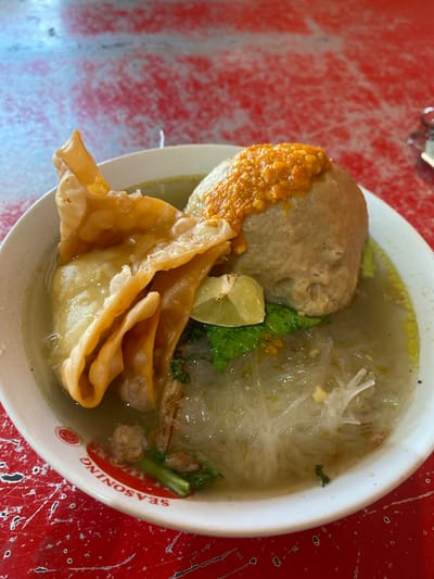 A close-up, overhead shot shows a bowl of Indonesian meatballs, known as "bakso," on a red, textured table. The white bowl has red lettering around the rim that reads "SEASONING" in reverse. Inside the bowl, a large, round meatball is topped with a dollop of orange chili sauce. To the left of the meatball, a golden-brown fried wonton is folded over. The broth contains clear glass noodles, chopped green onions, and small pieces of minced meat. A wedge of lime sits on top of the wonton. The table surface is predominantly red with white and black speckles, suggesting a worn or distressed finish. There are no people visible in the image.