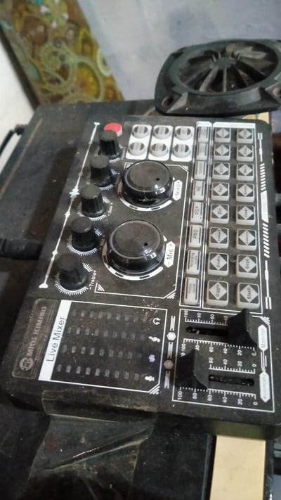 The image shows a close-up, high-angle shot of a black audio mixer with various knobs, buttons, and sliders. The mixer is dusty and appears to be used. The brand "MICO KIMISO" is visible on the left side, along with the text "Live Mixer". Several buttons are labeled with effects like "Reverb+", "Mic +", "Bass +", "Mid +", and "Treble+". To the right of these knobs are rows of square buttons, some of which have text such as "Karaoke", "MC", "Yell", "Din", "Chat", "Pop", "Pro", "Dindin", "Awesome", "Guys", "Primal Sound", "Giggle", and "Slash".
In the upper right portion of the image, a circular black fan is partially visible. Below the mixer, there are three sliders with numerical markings from 0 to 100, labeled "Monitor" and "Accompaniment". The background is blurred and appears to be an interior setting with patterned fabric on the left and a muted wall on the right. The lighting suggests indoor ambient light. The image is not specific to Banyusri, Indonesia, and no specific location details are evident within the frame.